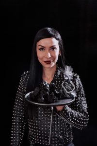 Katherine Sabbath's Christmas Coal collaboration with Kraken rum while holding a plate of black coal brownies in a leather jacket against a black background