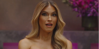 Chrishell Stause, host of Love Triangle, looks surprised while sitting in a warmly lit setting with vibrant purple flowers in the background